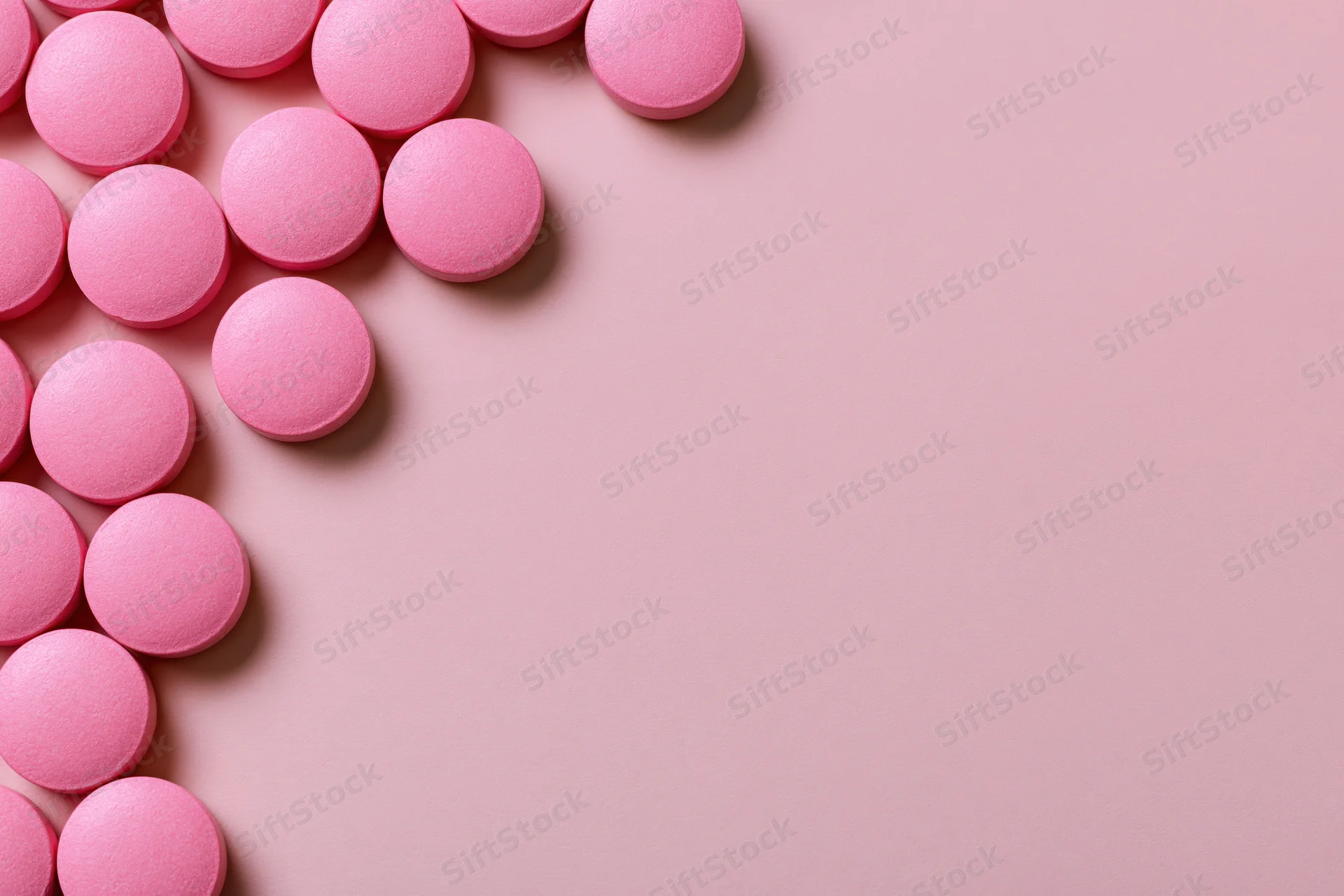 Scattered round pink supplement pills arranged loosely along the left edge on a pink surface with copyspace