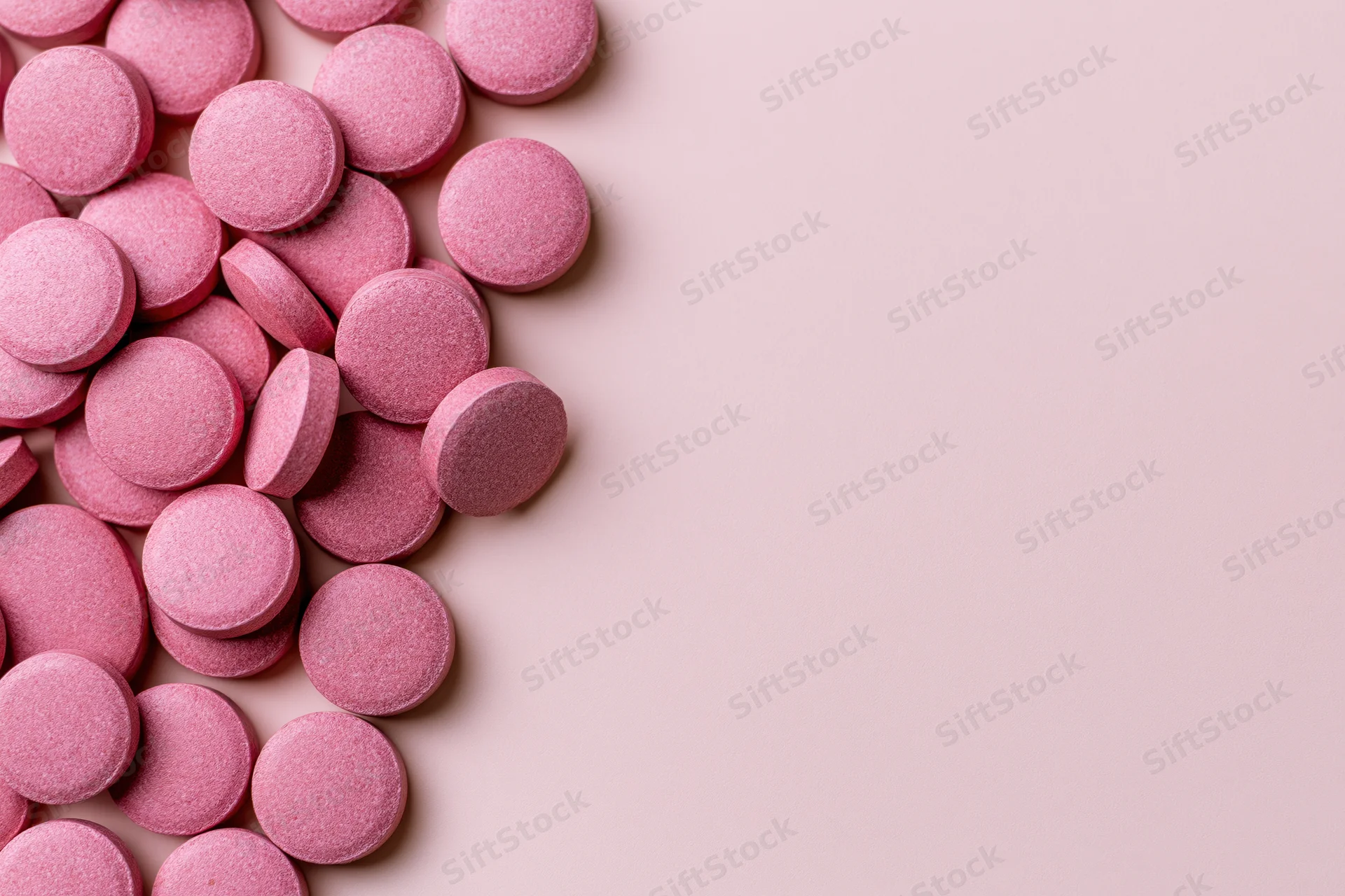 Scattered round pink supplement pills arranged loosely along the left side of a plain pastel pink surface with copyspace on the right