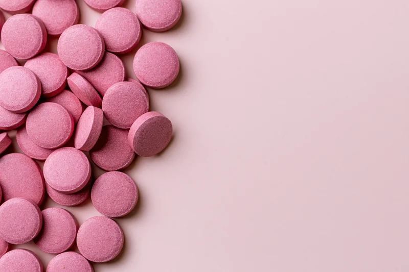Scattered round pink supplement pills arranged loosely along the left side of a plain pastel pink surface with copyspace on the right