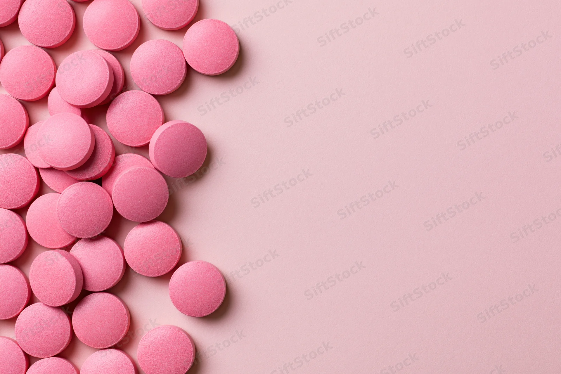 Scattered round pink supplement pills arranged loosely along the left side of a plain pink surface