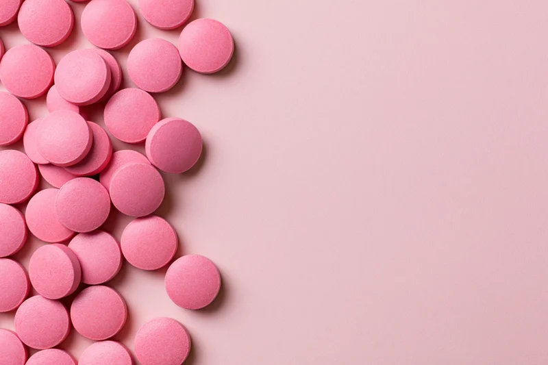 Scattered round pink supplement pills arranged loosely along the left side of a plain pink surface