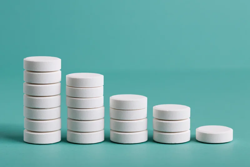 Stacks of white round pharmaceutical tablets arranged in descending order on a teal surface