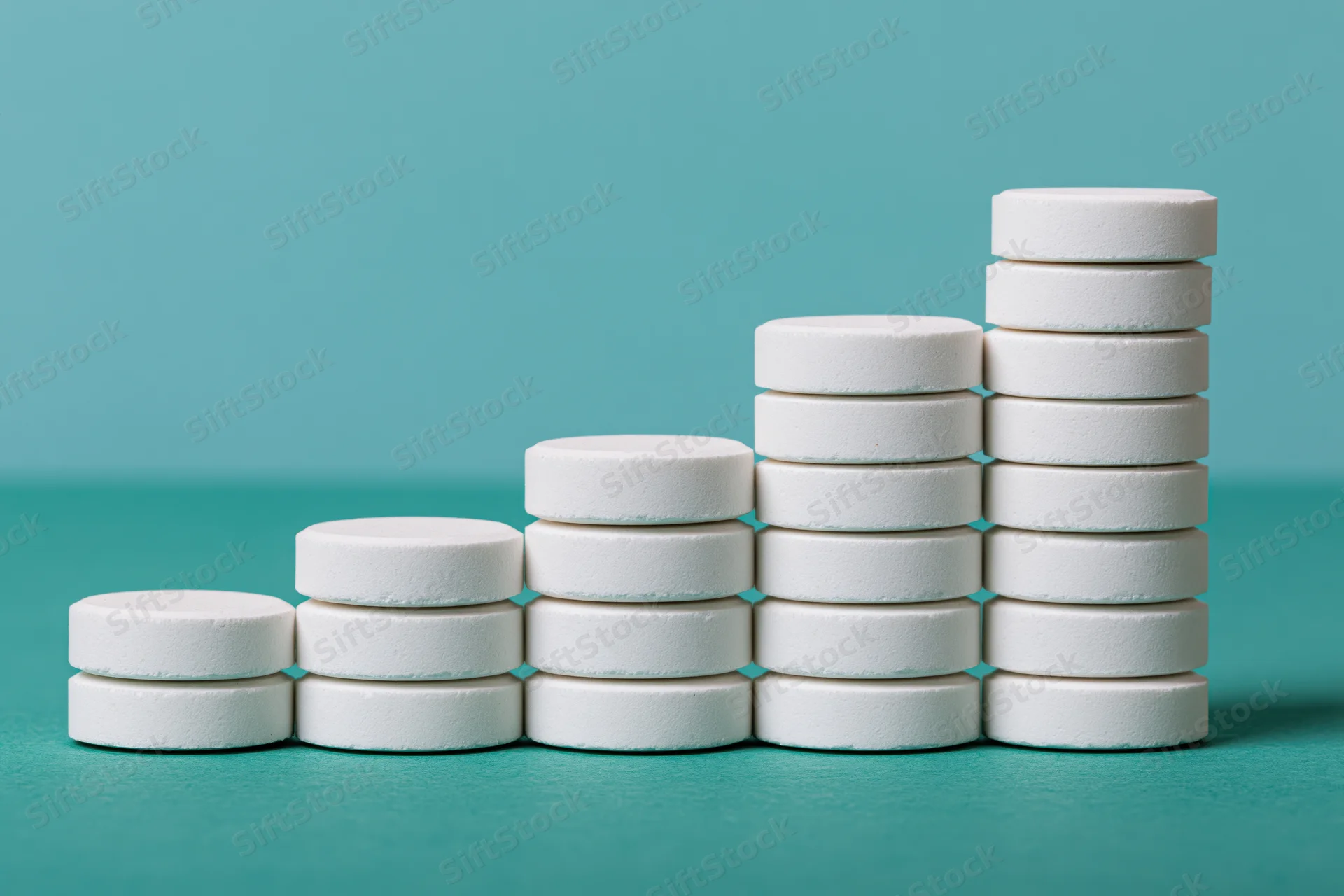 Stacks of white round pharmaceutical tablets arranged in ascending order on teal surface