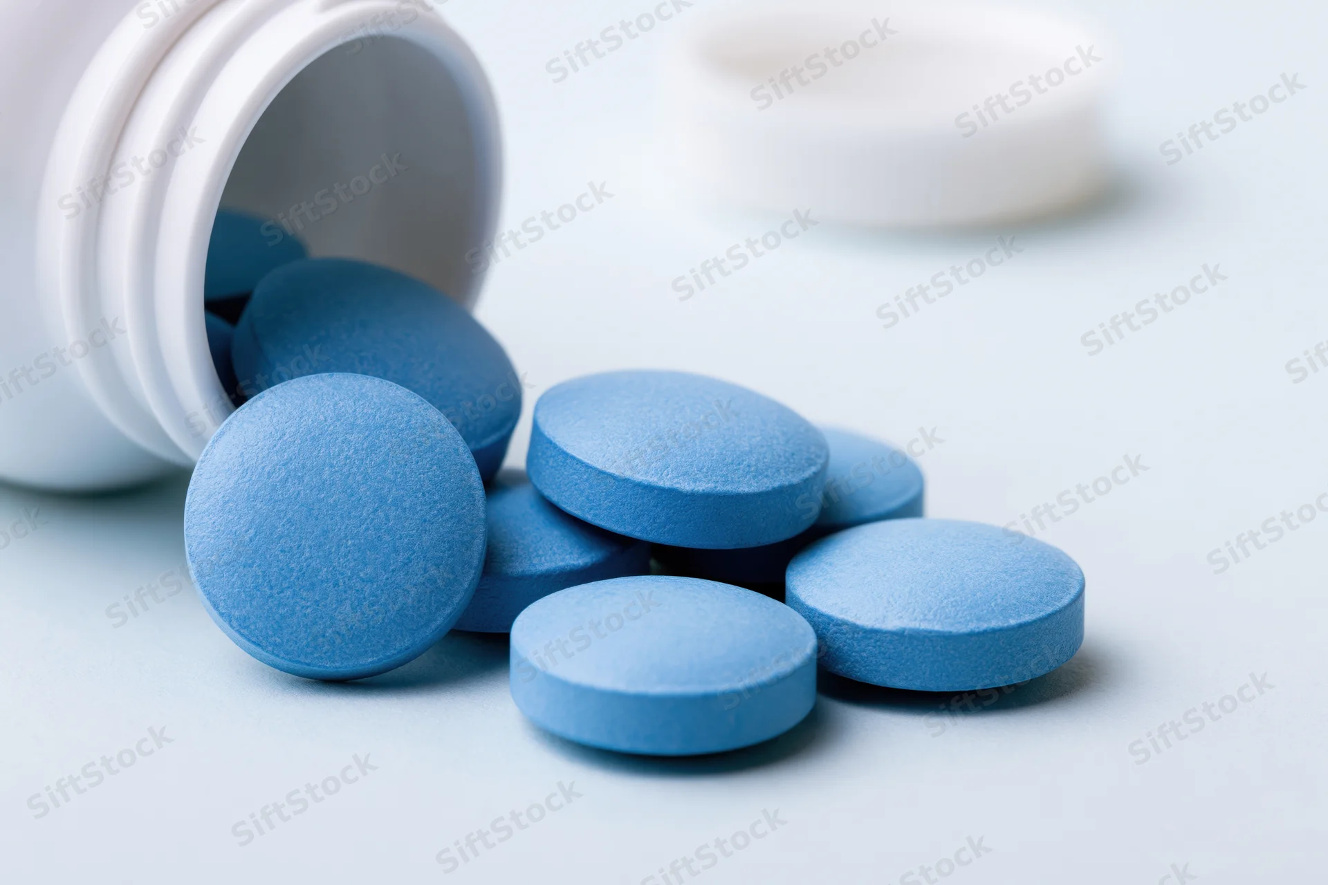 Blue pharmaceutical tablets spilled from a white bottle on a light surface