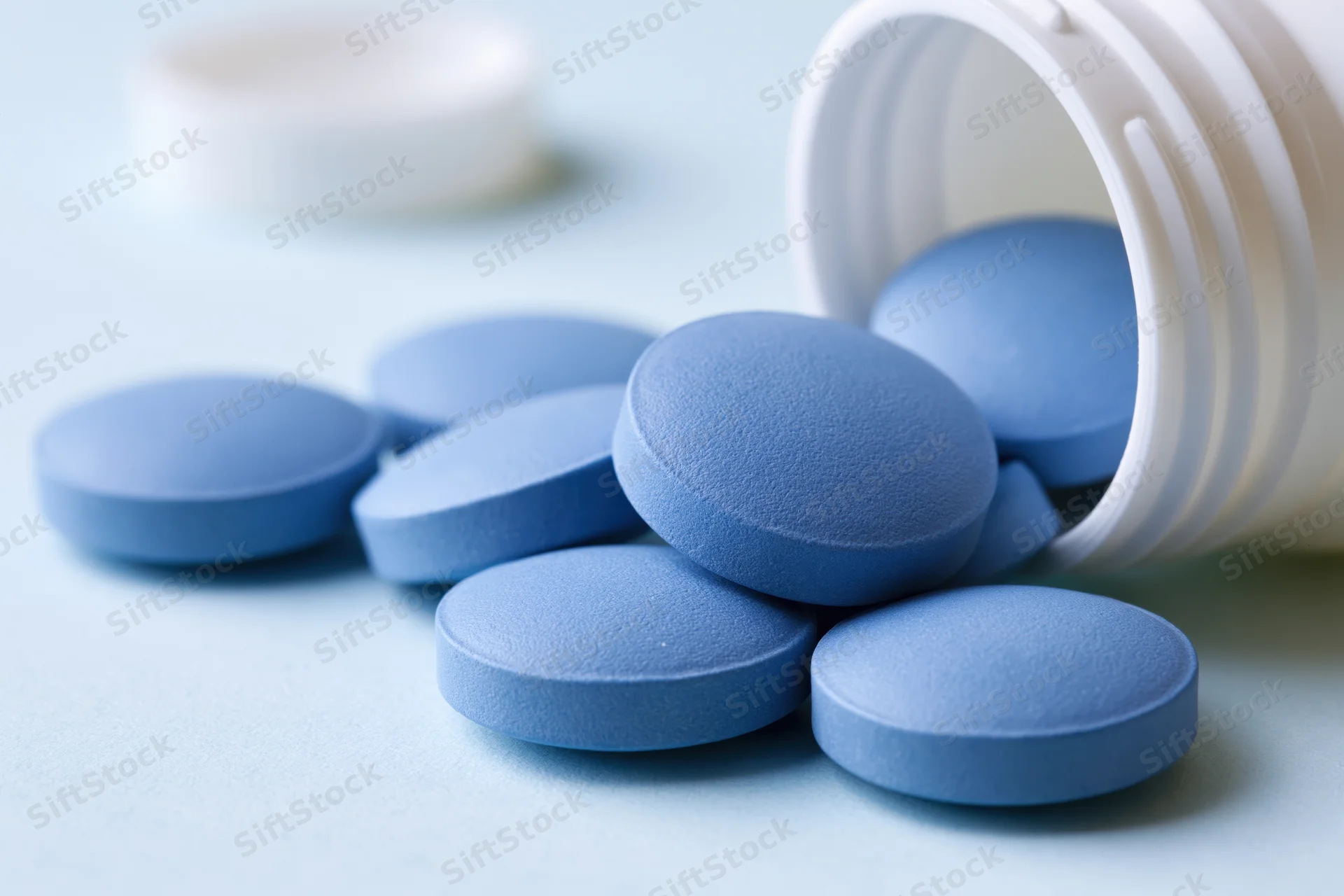 Blue pharmaceutical tablets spilled from a white plastic bottle on a light surface