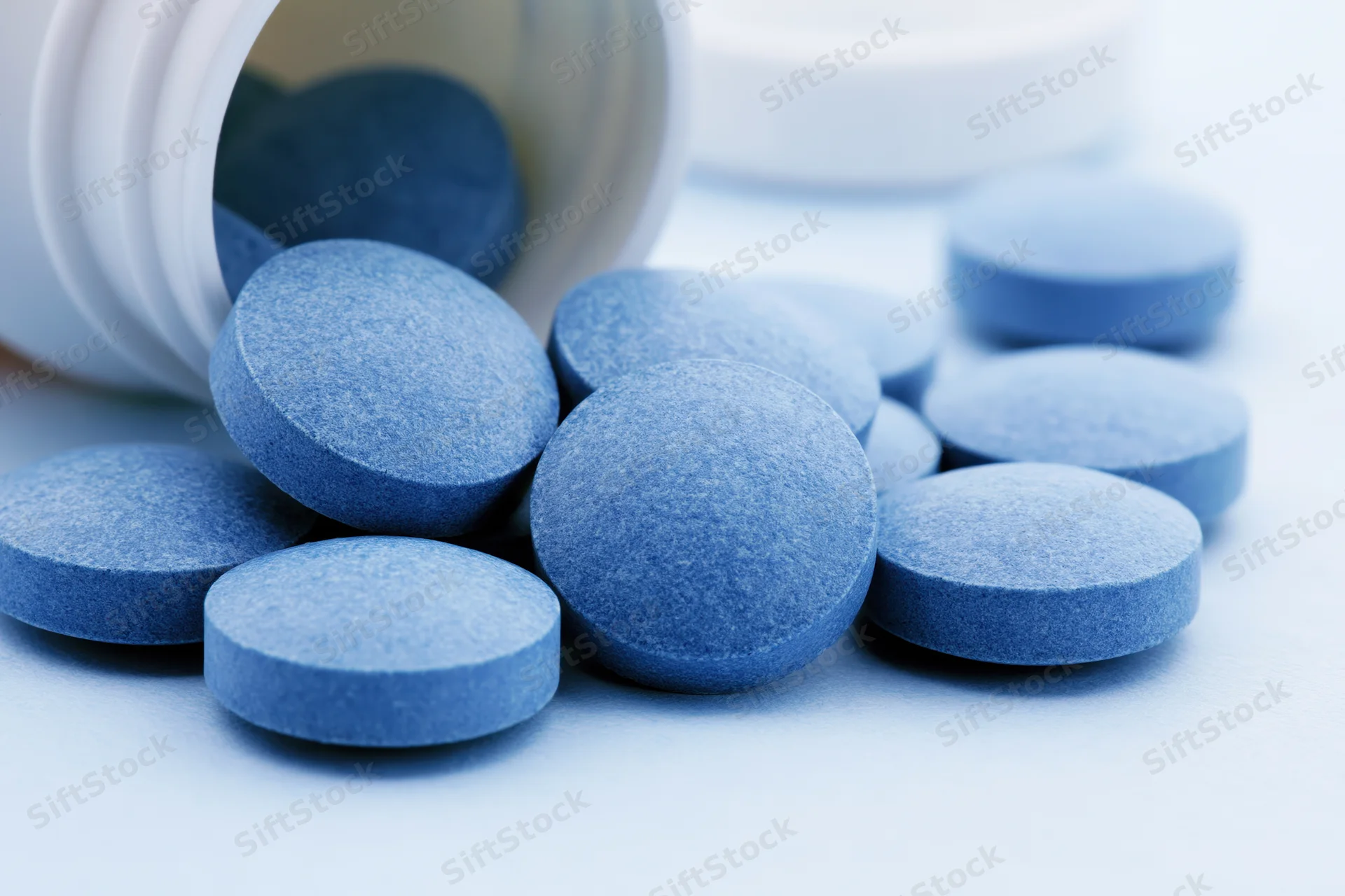 Blue pharmaceutical tablets spilled from a white bottle on a light surface