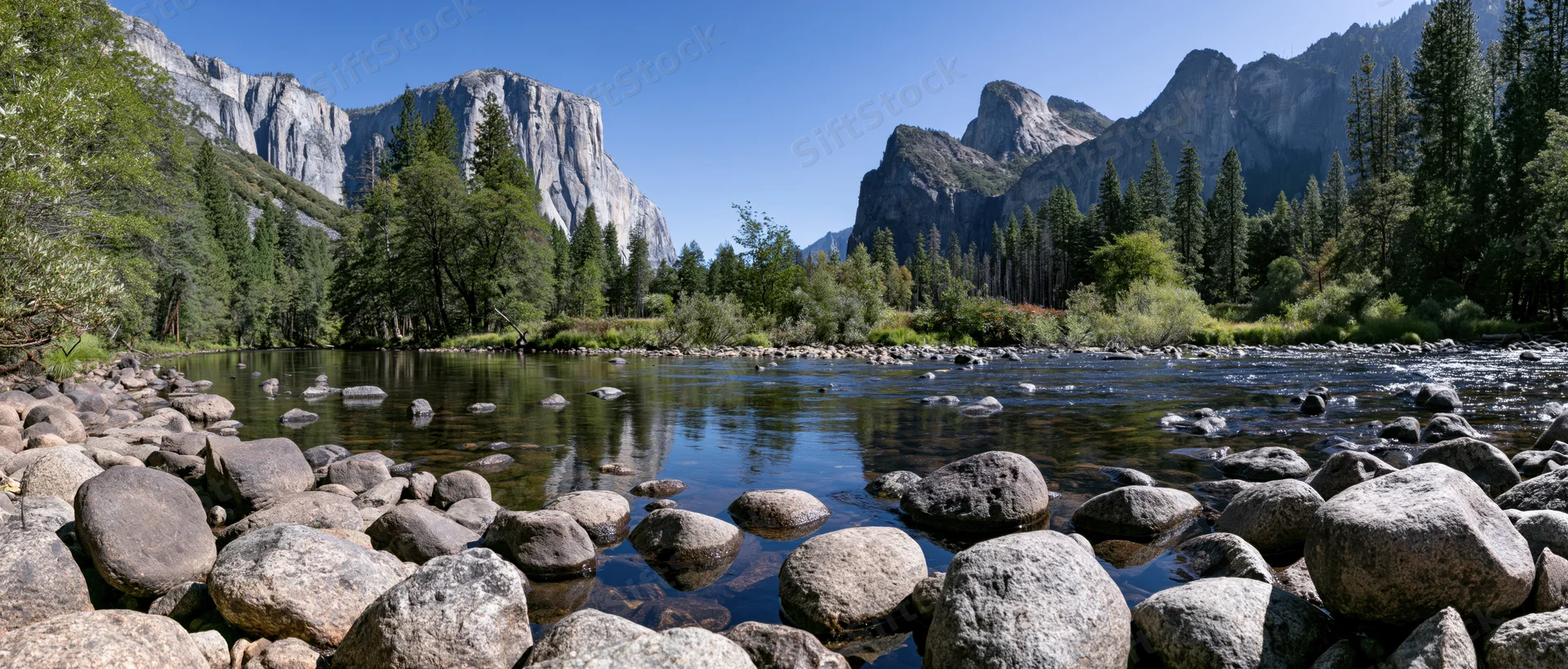 Shallow rocky river reflecting granite valley walls and dense forest under clear blue sky