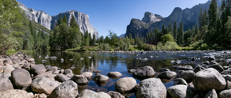 Shallow rocky river reflecting granite valley walls and dense forest under clear blue sky