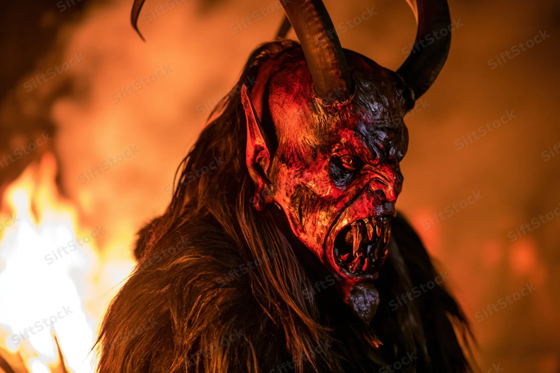 Krampus performer in carved wooden demon mask with horns and fangs, wearing fur costume, standing near fire at night
