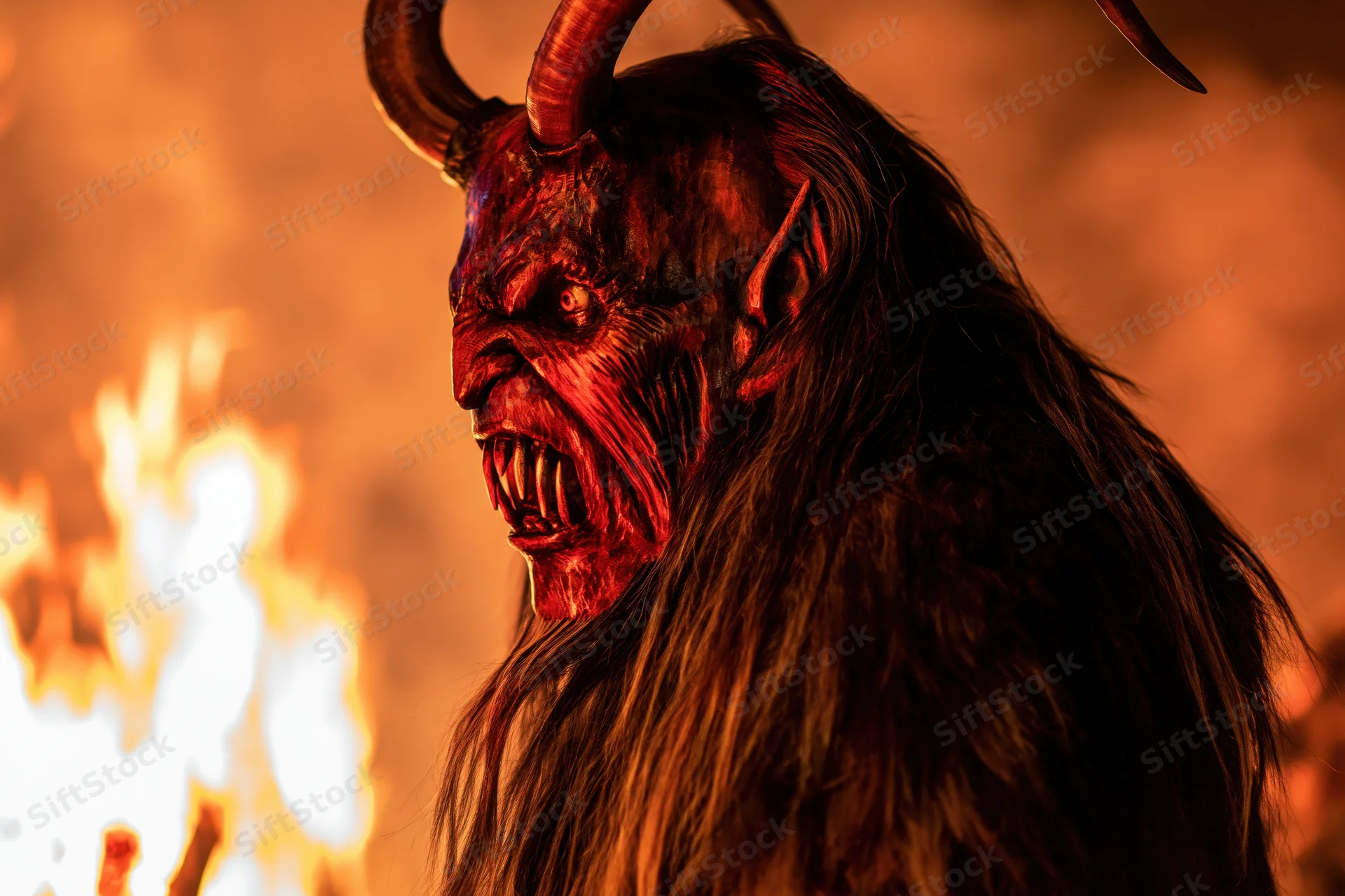 Krampus performer in carved wooden demon mask with horns and fangs, wearing fur costume, standing before flames at night