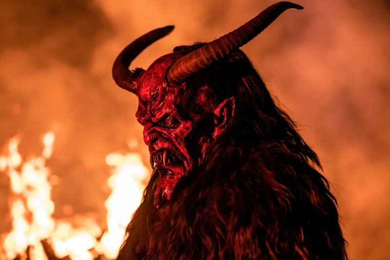 Krampus performer in wooden demon mask with horns and fur costume standing before flames at night
