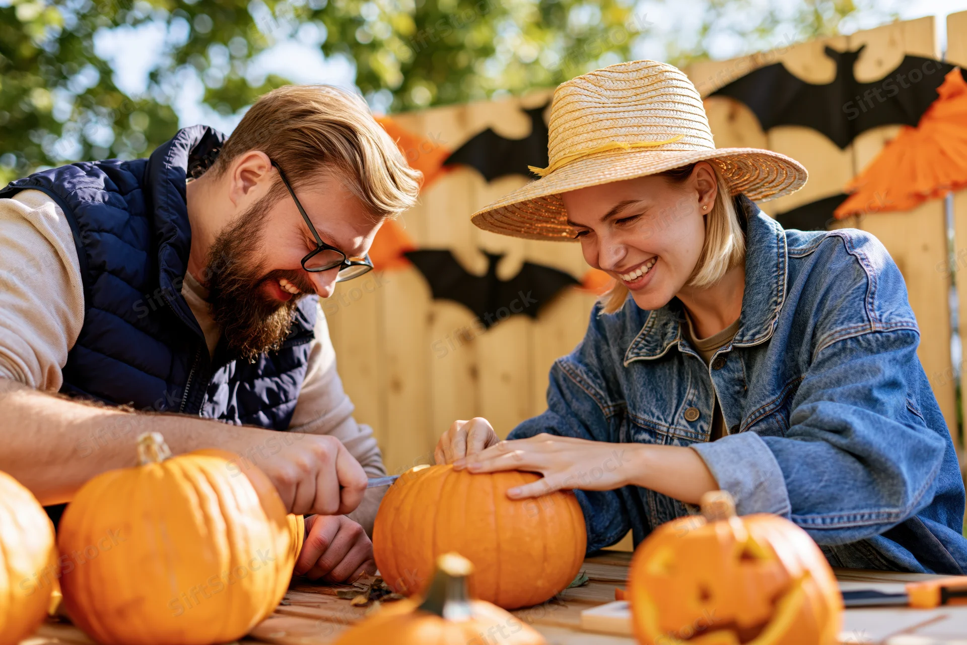 Couple carving pumpkins outdoors at a wooden table with halloween decorations in the background