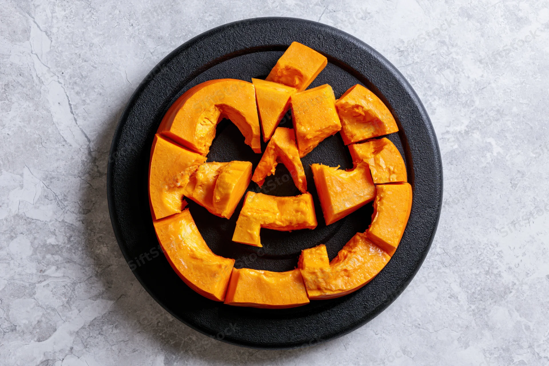 Carved pumpkin slices arranged in jack-o-lantern face on black circular plate on gray textured surface