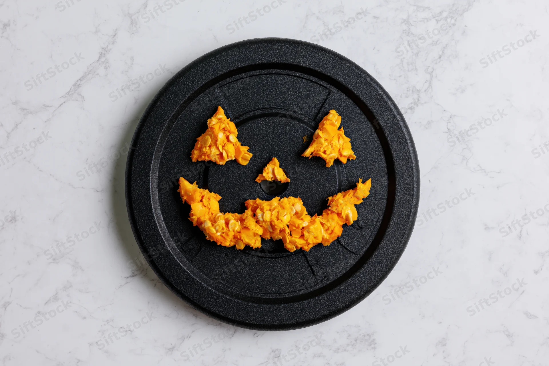 Jackolantern face made from orange snack pieces arranged on black gym weight plate on marble surface