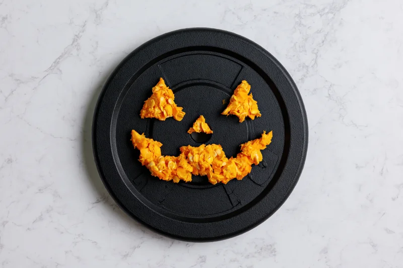 Jackolantern face made from orange snack pieces arranged on black gym weight plate on marble surface