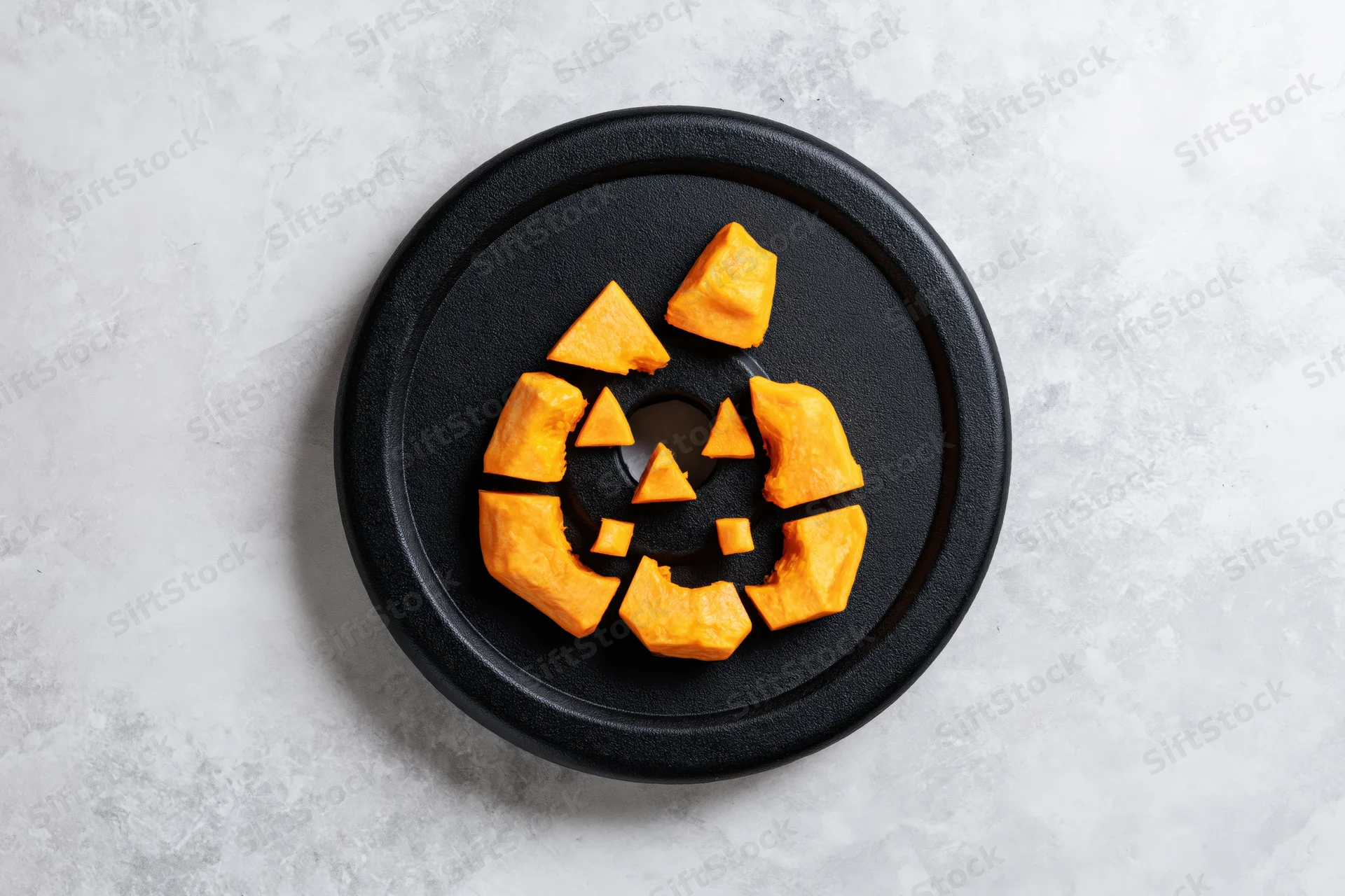Carved pumpkin pieces arranged as a jack-o-lantern face on a black gym weight plate