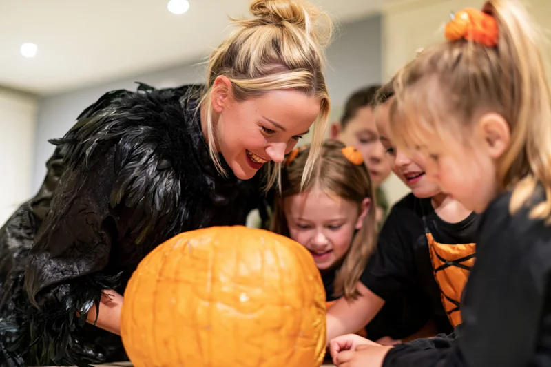 Woman in black feathered costume smiling while carving a pumpkin with children indoors
