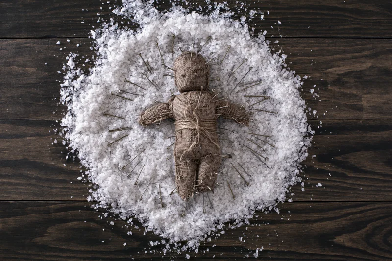 Voodoo doll made of burlap and twine, pierced with pins, lying on a circle of salt on dark wooden surface