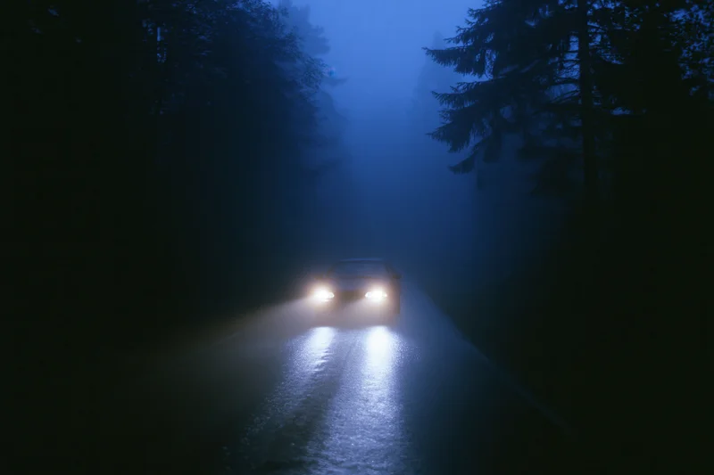 Car driving toward camera on narrow forest road at night with headlights cutting through fog and wet pavement