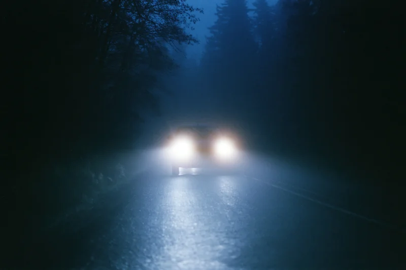 Car driving toward camera on narrow forest road at night with headlights cutting through fog and wet pavement