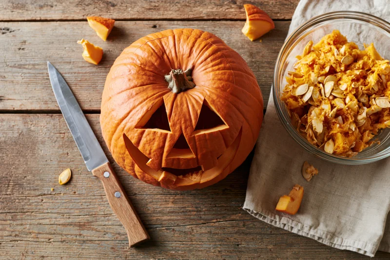Carved jack-o-lantern pumpkin with triangular face on wooden table beside knife and bowl of seeds