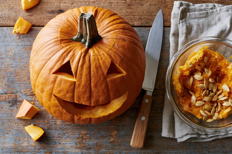 Carved jack-o-lantern pumpkin with triangular eyes and mouth on wooden table beside knife and bowl of seeds