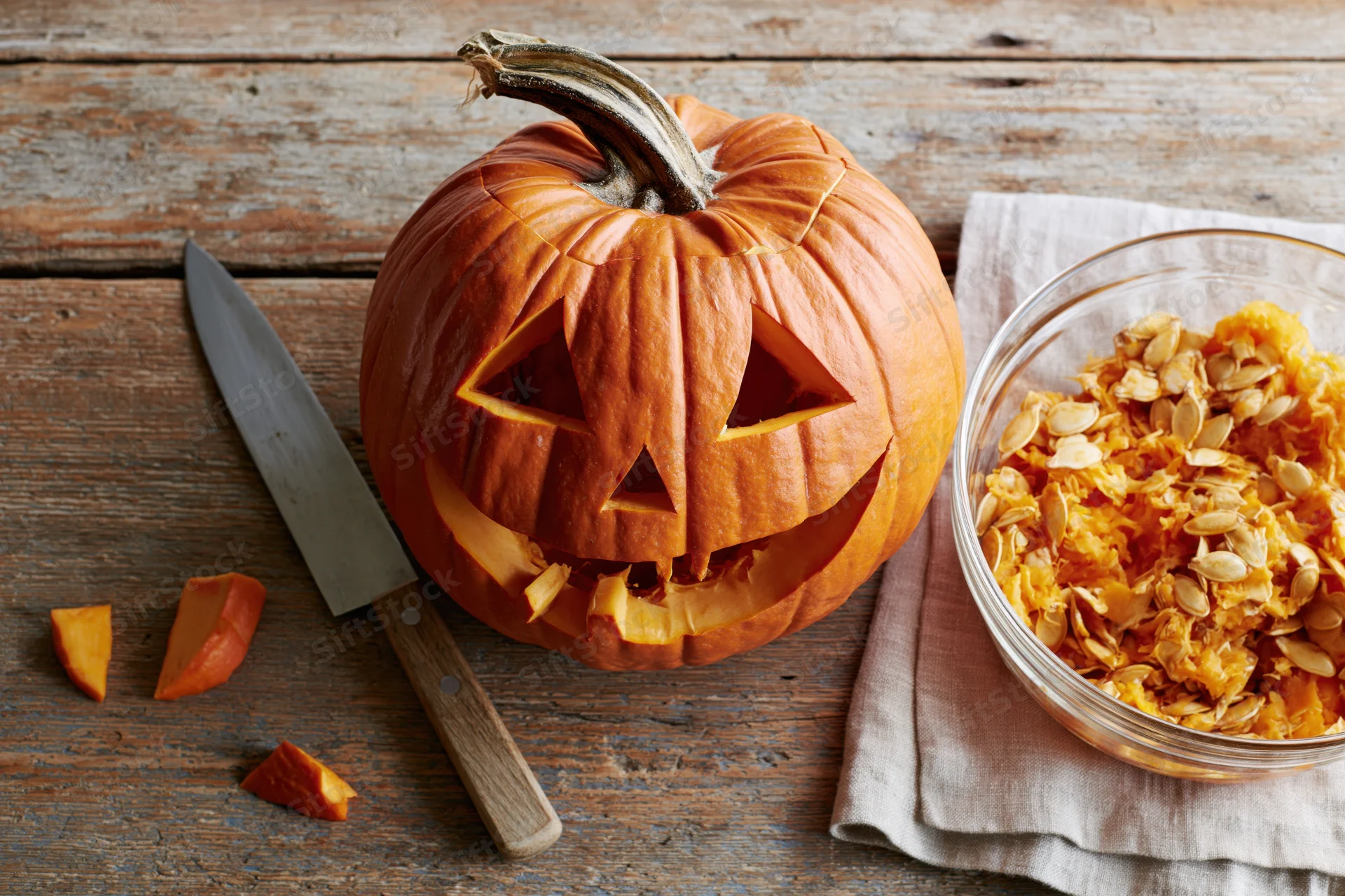 Carved jack-o-lantern pumpkin with triangular eyes and mouth on wooden table beside bowl of seeds and knife
