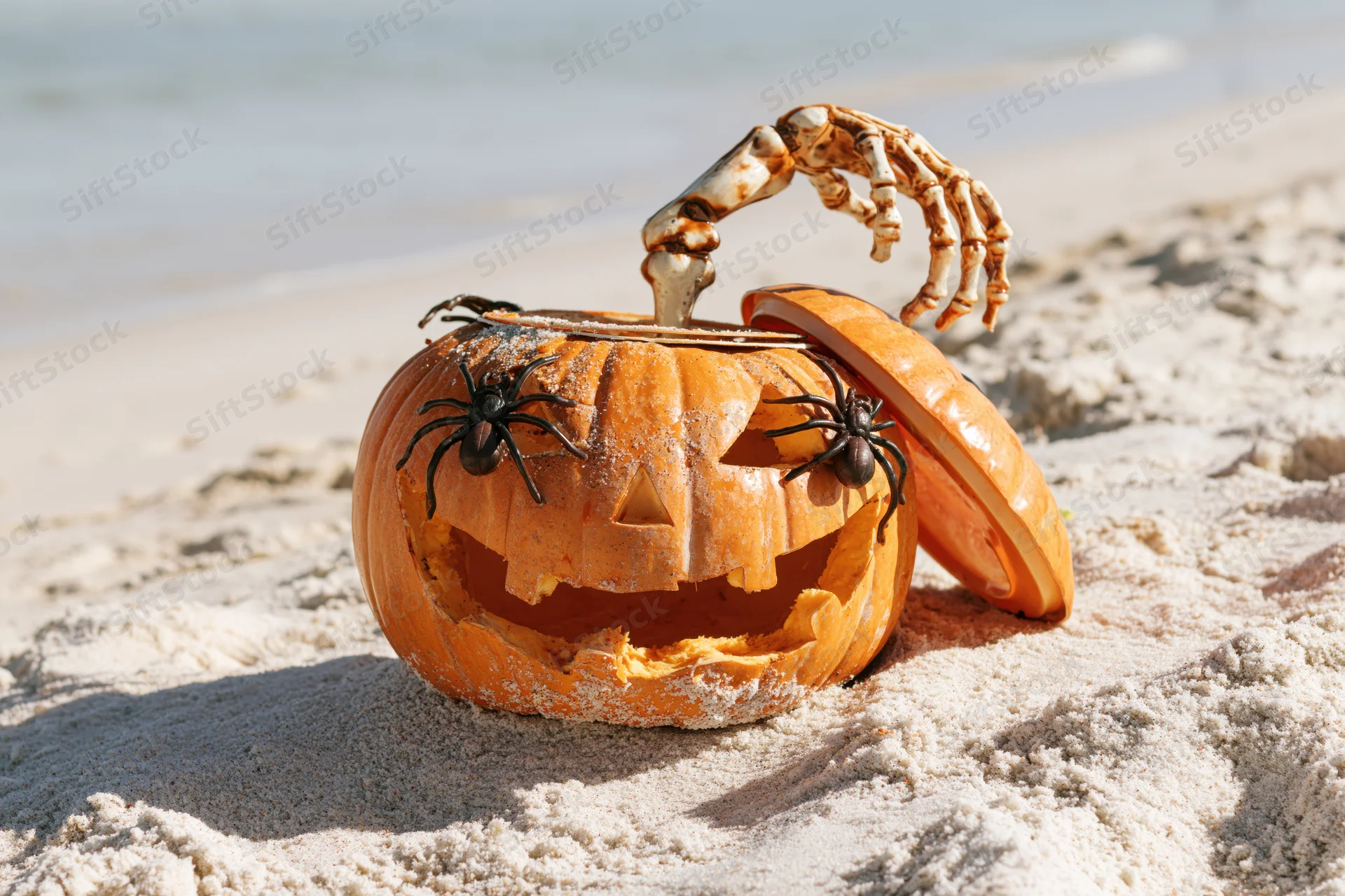 Carved jack-o-lantern with skeleton hand and spiders on sandy beach near ocean