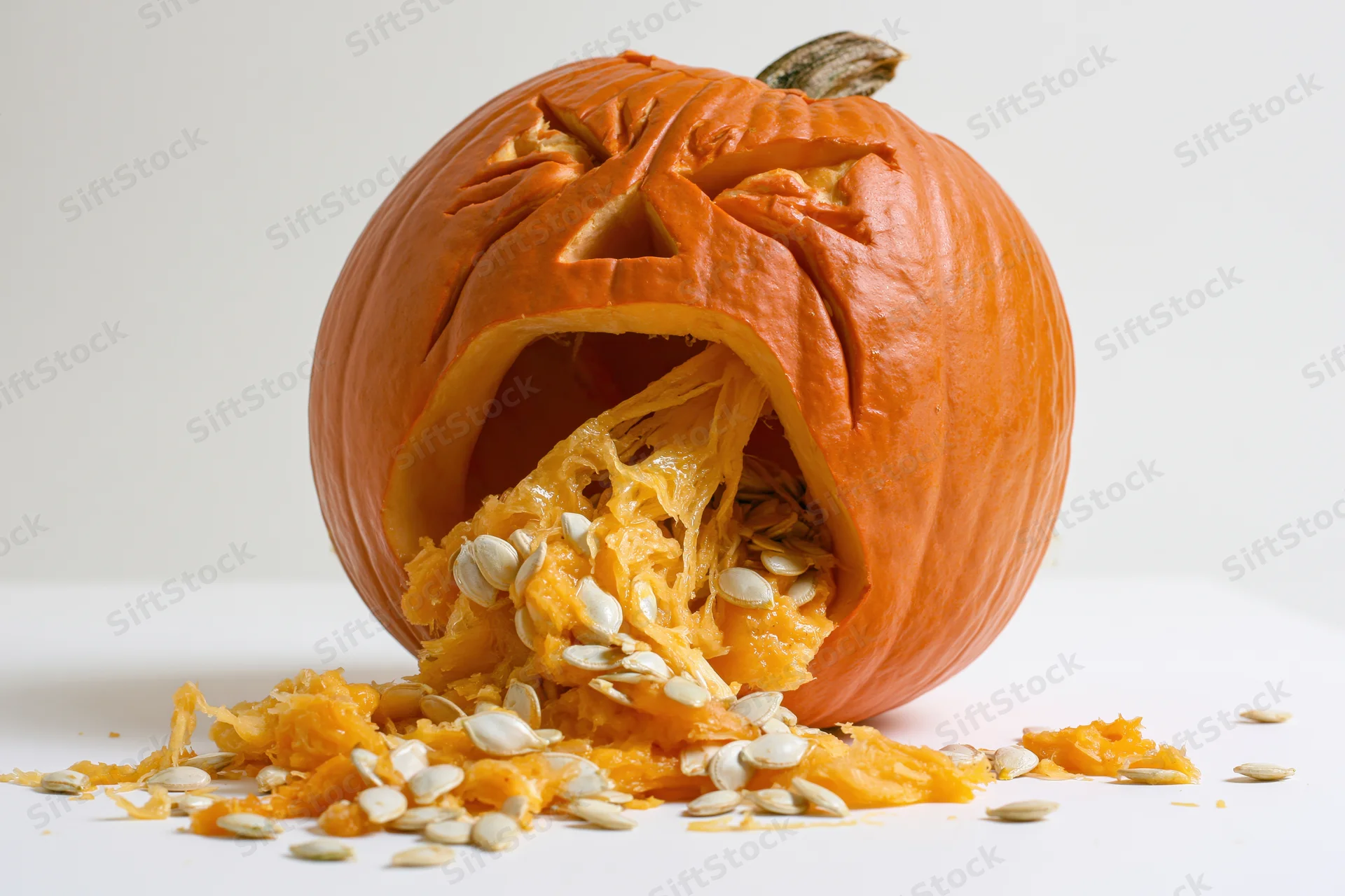 Carved pumpkin with anguished expression spilling seeds and pulp on white surface