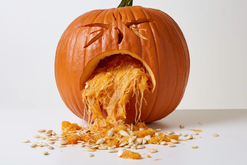 Carved pumpkin with anguished expression and exposed pulp and seeds on white surface