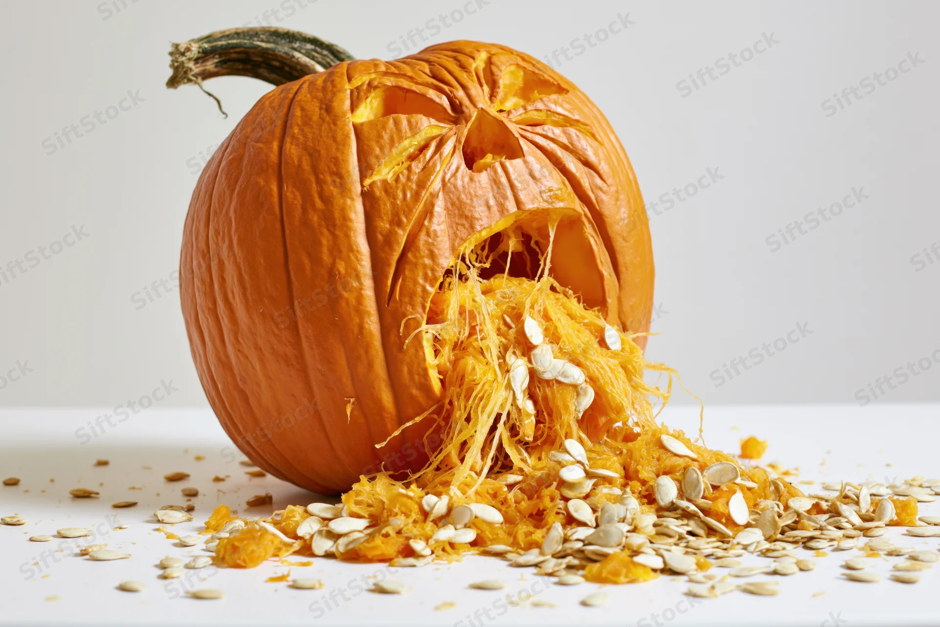 Carved pumpkin with anguished expression spilling seeds and pulp on white surface