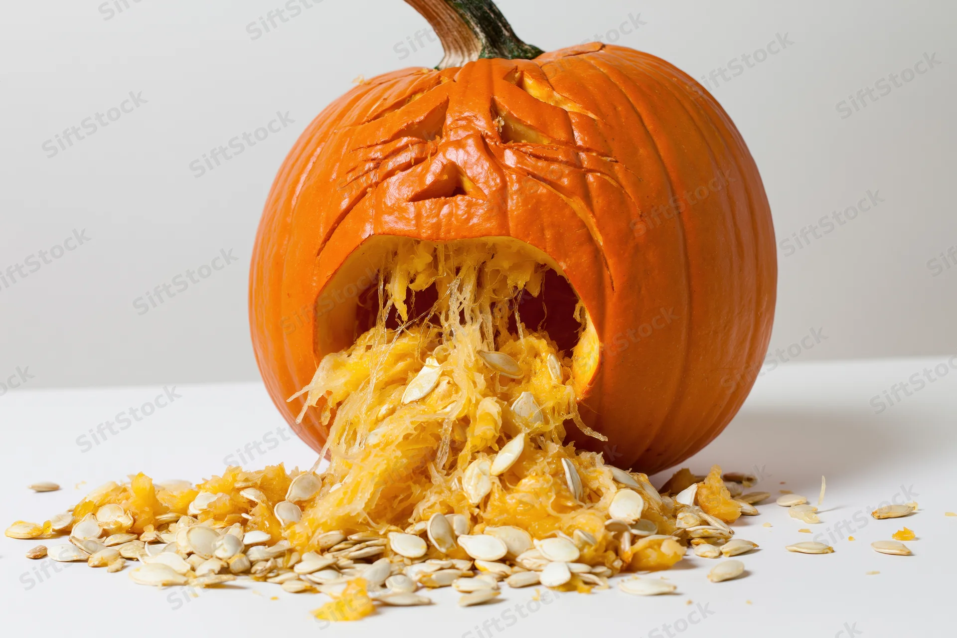 Carved pumpkin with anguished expression spilling seeds and pulp on white surface