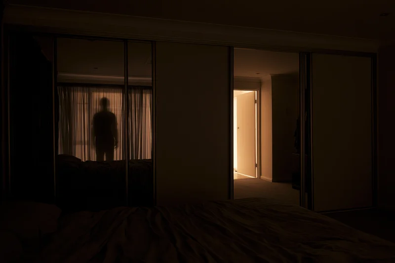 Dark bedroom at night with unmade bed in foreground and silhouette reflected in mirror