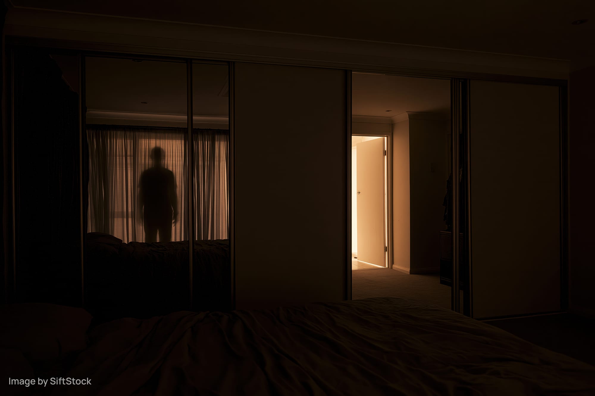 Dark bedroom at night with unmade bed in foreground and silhouette reflected in mirror