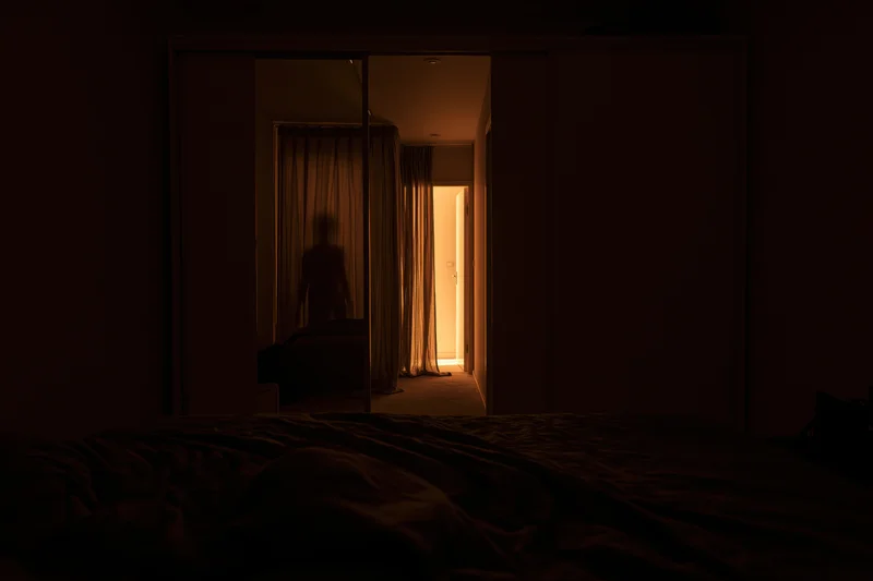 Dark bedroom at night with unmade bed in foreground and silhouette visible in hallway doorway