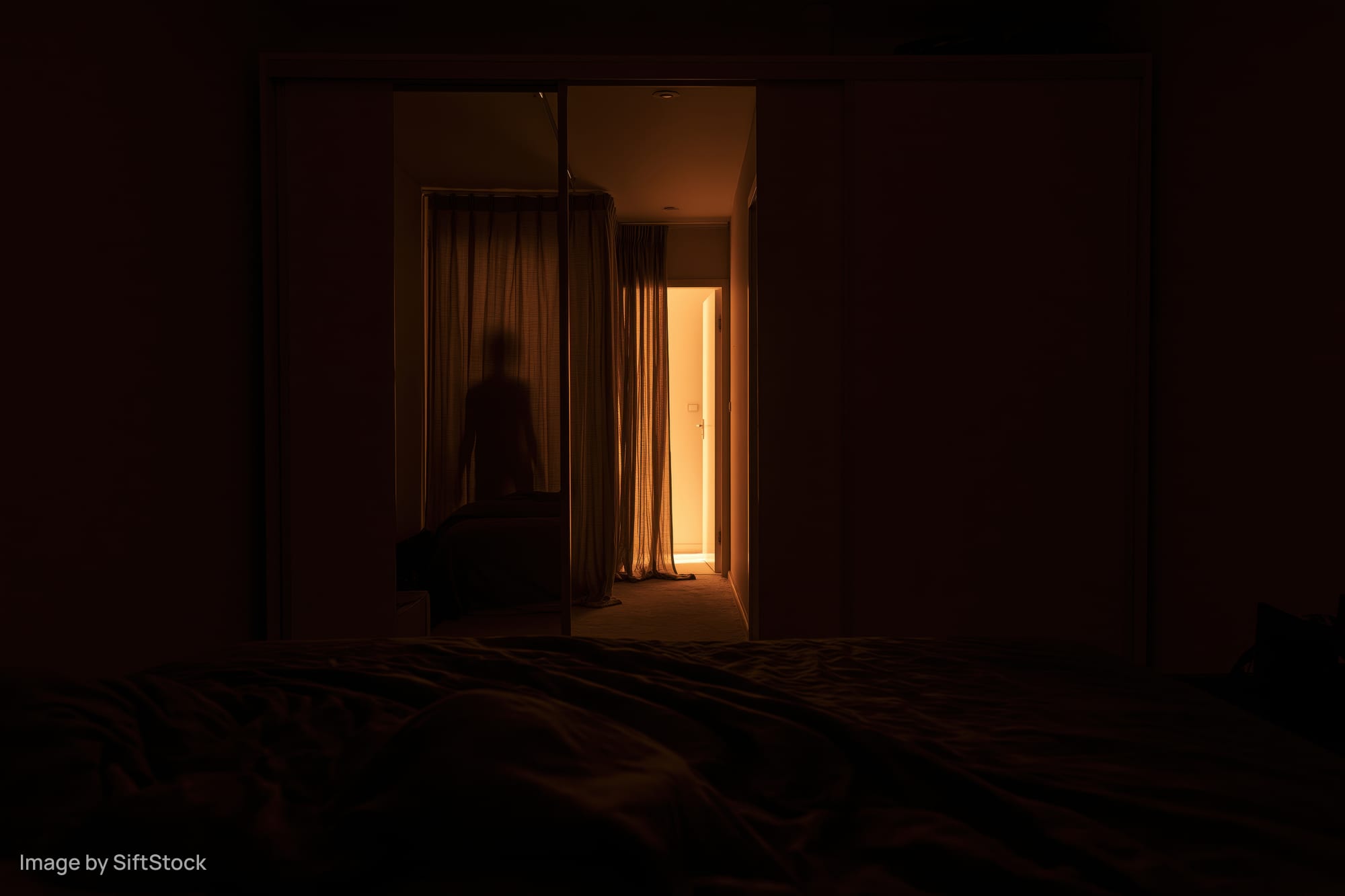 Dark bedroom at night with unmade bed in foreground and silhouette visible in hallway doorway
