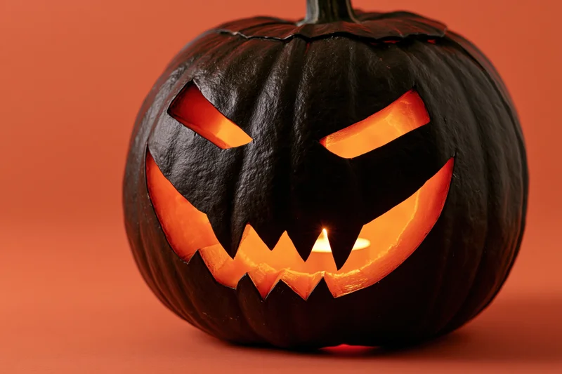 Dark carved jack-o-lantern with jagged smile and glowing eyes on orange background