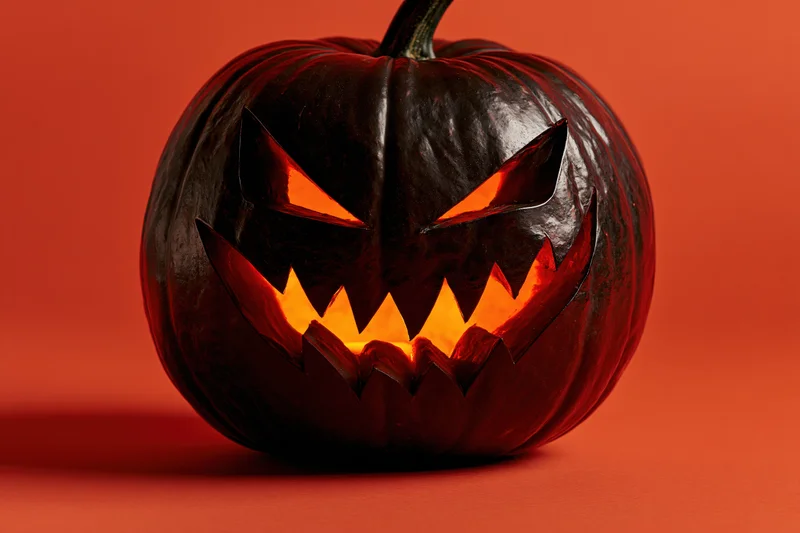 Dark carved jack-o-lantern with glowing jagged teeth and eyes on orange background
