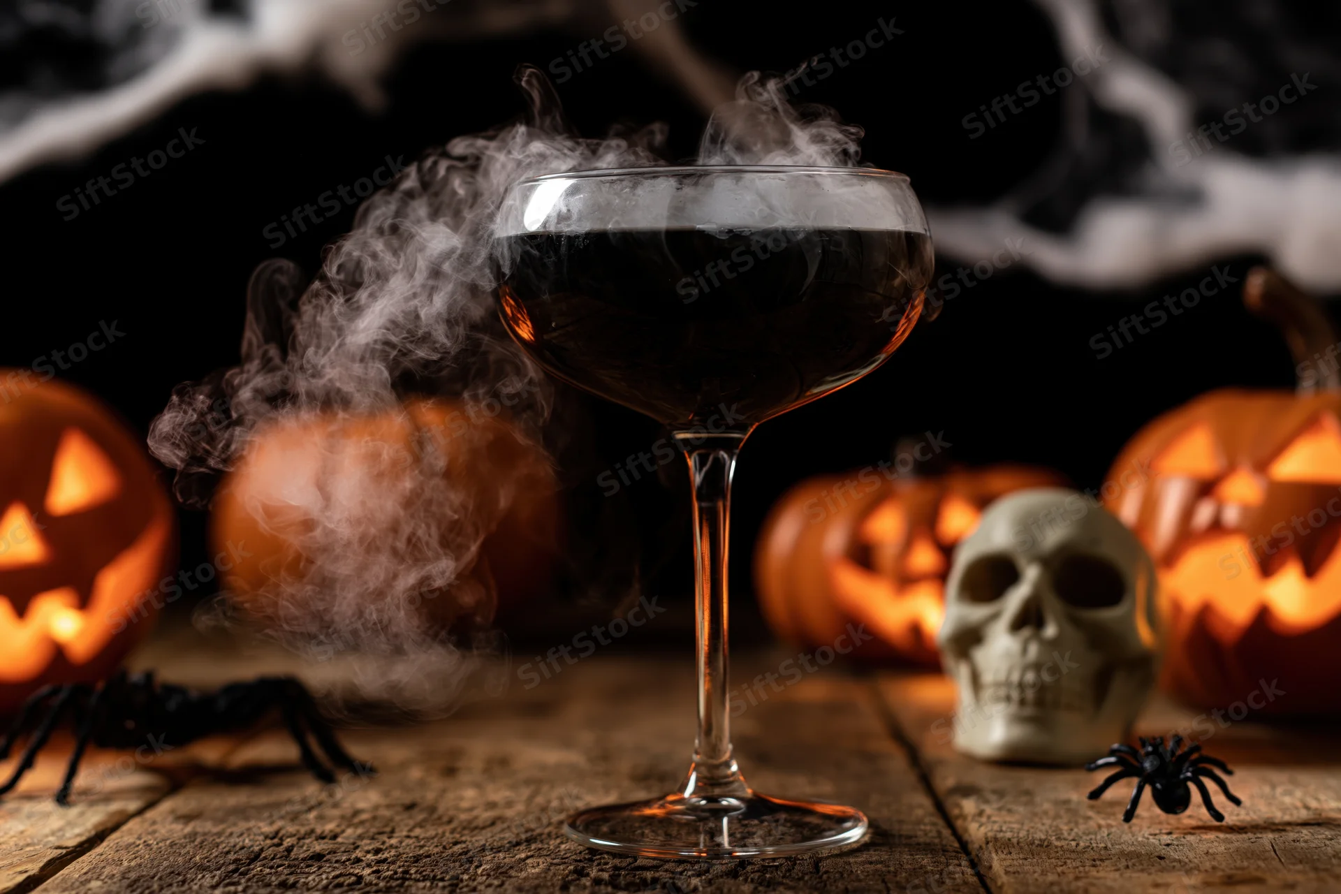 Dark cocktail glass with smoking black liquid on wooden table surrounded by carved pumpkins, skull, and spiders for halloween decoration