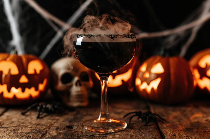 Black cocktail in a coupe glass with smoke, surrounded by glowing jack-o'-lanterns and Halloween decorations on a wooden table