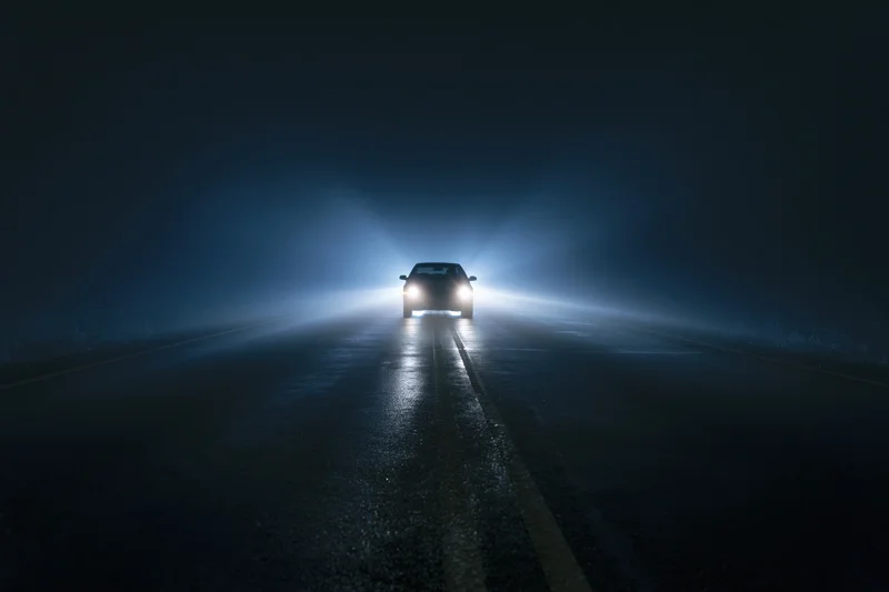 Dark sedan driving toward camera on empty two-lane road at night with headlights on
