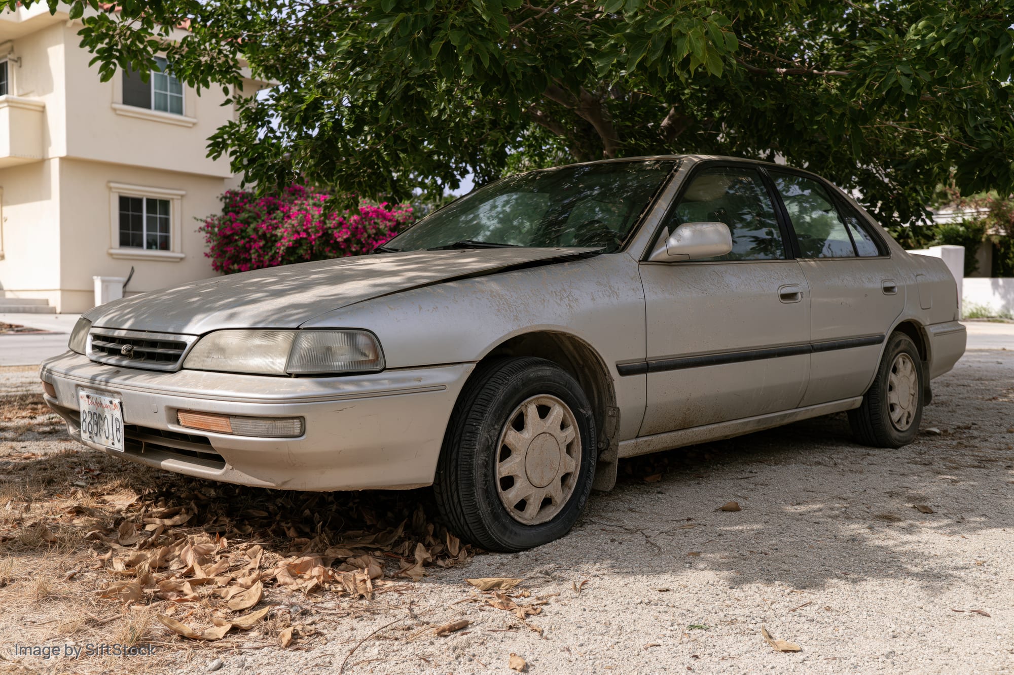 Dusty silver mid-1990s japanese sedan parked on dry gravel under tree shade near residential house