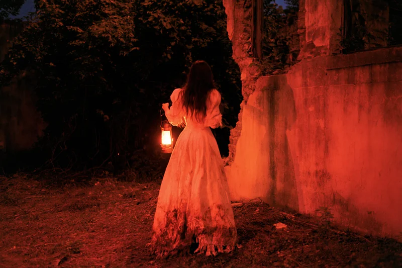 Woman in tattered victorian gown holding lantern walking through overgrown ruins at night