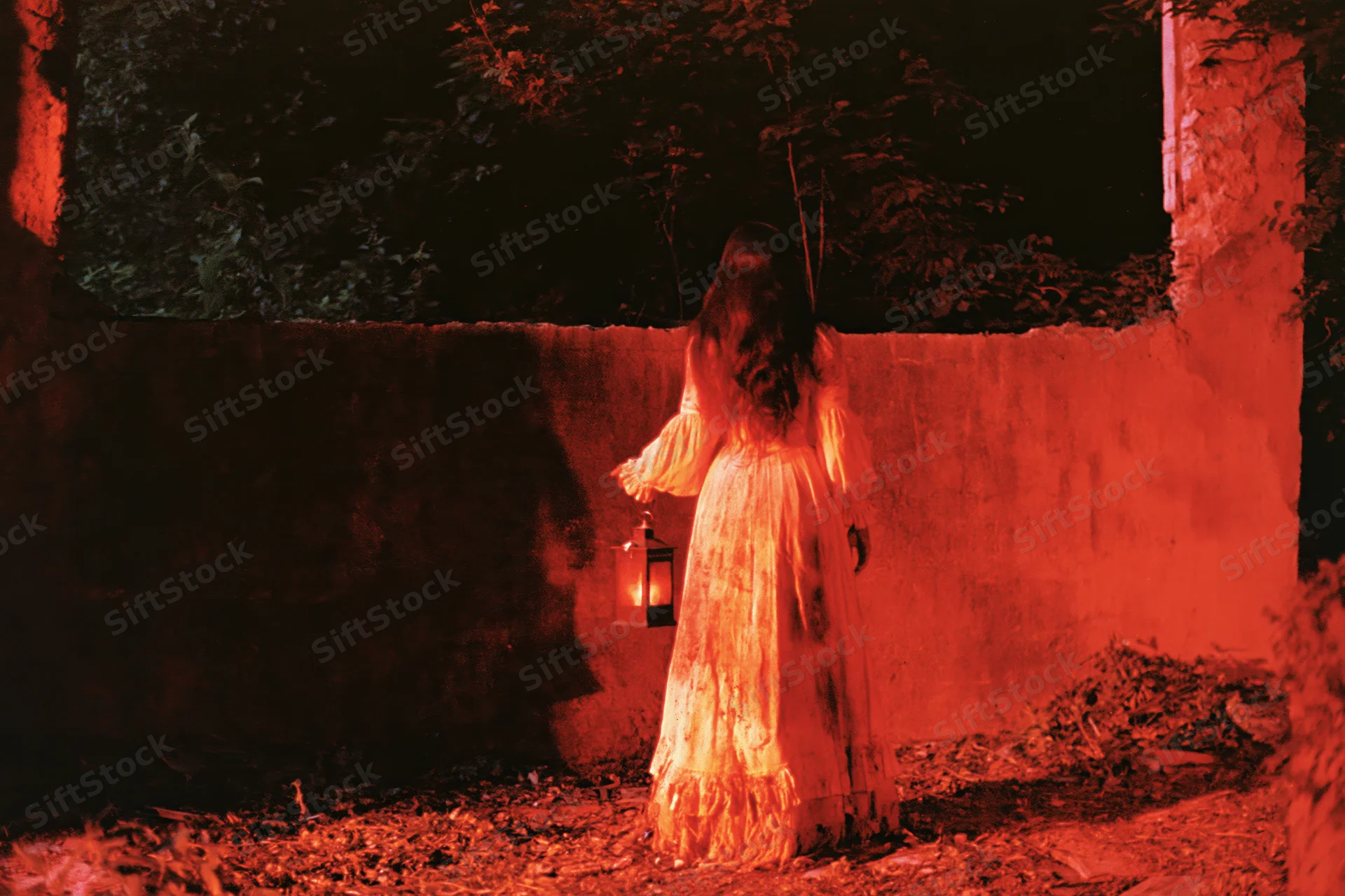 Woman in tattered victorian gown holding lantern facing away from camera in dimly lit garden at night
