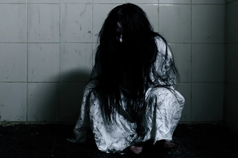 Woman in white gown crouching in dark tiled corner of asylum
