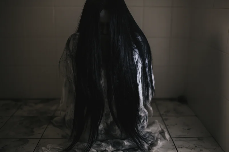 Woman with long black hair covering face sits in dark tiled corner of bathroom