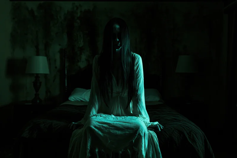 Ghostly woman in white gown sitting on bed in dark bedroom with green light