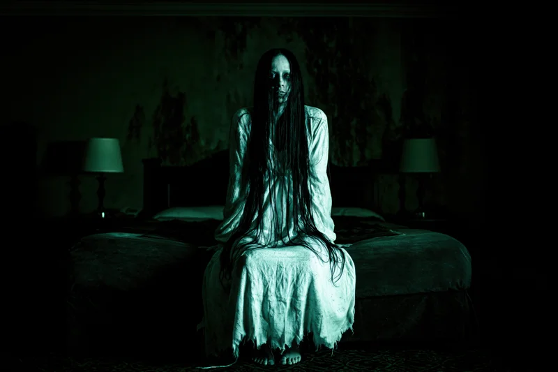 Ghostly woman with long dark hair sitting on bed in dimly lit bedroom
