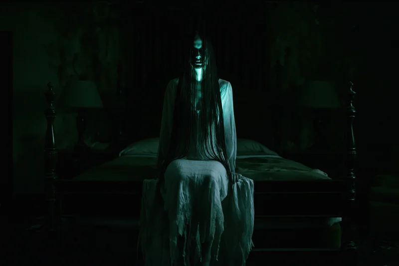 Ghostly woman with long dark hair sitting on bed in dimly lit bedroom