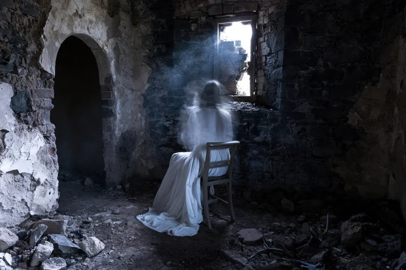 Translucent ghostly figure seated on wooden chair in dark ruined stone interior with light from window